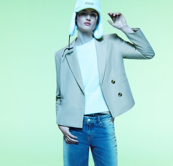 Beżowa marynarka dwurzędowa z złotymi guzikami, biała sweter, niebieskie dżinsy i jasnoniebieska czapka z uszami, na zielonym tle.