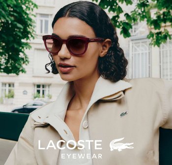 Gafas de sol con montura burdeos, forma de ojo de gato redondeada y lentes oscuros. Modelo vistiendo una chaqueta beige con cuello blanco y el logo de Lacoste.