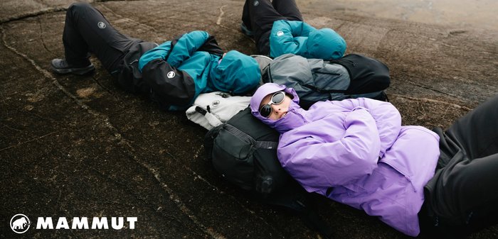 Vier Personen liegen auf einer felsigen Oberfläche und tragen Jacken in Türkis, Grau und Lila. Eine Person trägt Sonnenbrillen und eine lila Jacke.