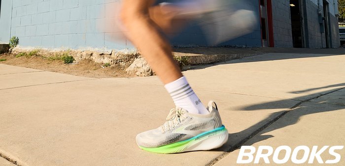 Lichtgrijze hardloopschoen met een groene verloopzool, een gestructureerd oppervlak en minimale vetering, gedragen met wit-grijs gestreepte sokken.