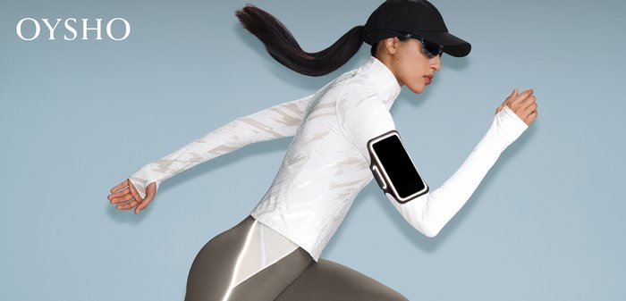 Frau in weißem Langarmshirt, grauer Leggings mit reflektierendem Streifen, schwarzer Mütze und Sonnenbrille, die mit einem Handy-Armband vor einem hellblauen Hintergrund läuft.