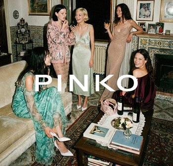 Five women dressed in elegant evening wear socialize in a cozy living room with wine and books on a wooden coffee table.