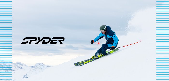 Figure de ski portant une veste isolante bleue et noire, un pantalon sombre et un casque. Skis verts avec des accents vifs sur un terrain recouvert de neige.