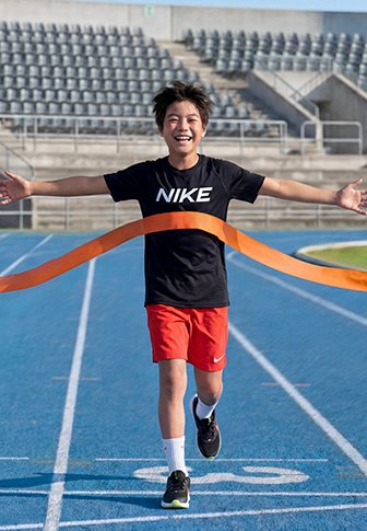 Băiat în tricou Nike și pantaloni scurți roșii trece fericit linia de sosire pe o pistă de alergare albastră, cu scaune goale în tribună în fundal.
