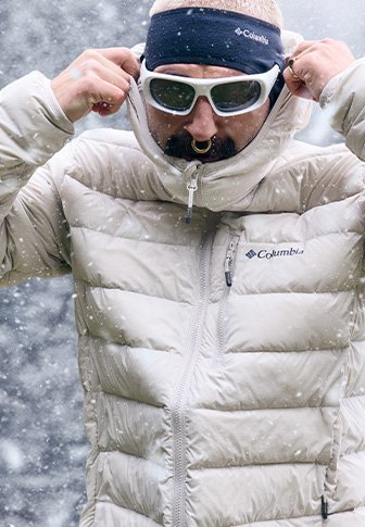 Φουσκωτό μπεζ μπουφάν με ψηλό γιακά και κλείσιμο με φερμουάρ. Το άτομο φορά λευκά γυαλιά ηλίου, μαύρη κορδέλα και βρίσκεται σε ένα χιονισμένο περιβάλλον.