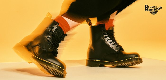 Zwarte leren Dr. Martens-laarzen met gele stiksels, gedragen met oranje sokken, tegen een gele achtergrond tijdens een halverwege stap.