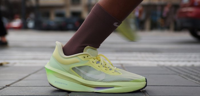 Άτομο που φοράει ελαφρύ πράσινο παπούτσι για τρέξιμο Nike ZoomX με μοβ κάλτσες, πατάει στο αστικό πεζοδρόμιο κοντά σε διάβαση πεζών.