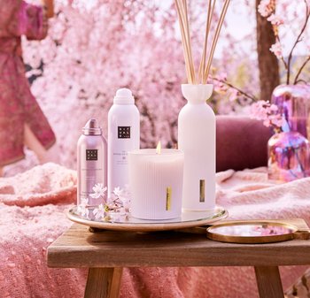 Hvid ribbet lys med gulddetalje, lyserøde sprayflasker og en hvid vase på et træbakke, mod en blød lyserød baggrund.