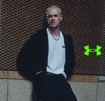 Jonge man met kort blond haar, gekleed in een zwart jack, wit T-shirt en zwarte broek, staand tegen een metalen gaaswand met een groen logo.