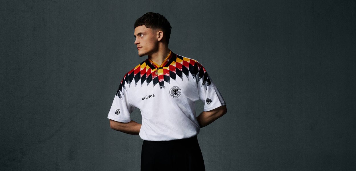 Junger Mann in einem Vintage Fußballtrikot Deutschlands mit geometrischem roten, gelben und schwarzen Muster, der mit den Händen hinter dem Rücken vor dunklem Hintergrund steht.
