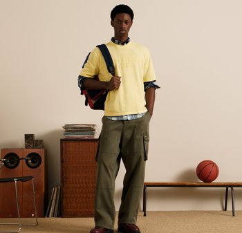 Žltá bavlnená grafická mikina s krátkymi rukávmi, olivové cargo nohavice a modro-červený batoh. Stojí na béžovom pozadí s basketbalom.