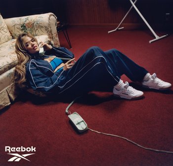 Mujer con chándal azul y zapatillas blancas yace en un suelo alfombrado en rojo, sosteniendo un teléfono con cable cerca de un sofá floral en una habitación con poca luz.