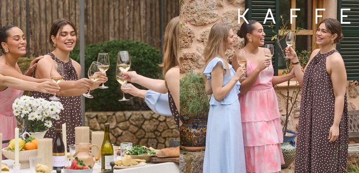 Drei Frauen in Sommerkleidern lächeln und stoßen mit Weingläsern im Freien nahe einer Steinmauer und grünen Fensterläden an.