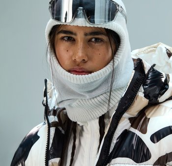 Kurtka puchowa z błyszczącym czarno-białym wzorem w kamuflaż, zestawiona z jasnoszarym wełnianym szalikiem i sportowymi okularami przeciwsłonecznymi.