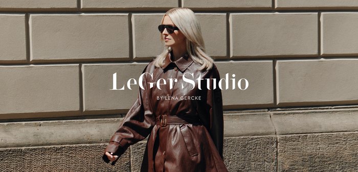 Abito camicia in pelle marrone con vita cintata. La modella indossa collant neri trasparenti e tacchi alti, mentre cammina contro un muro di pietra testurizzato.