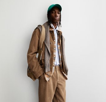 Junger Mann in geschichteten braunen und beigen Cord-Outfits, mit grüner Mütze und einer beigen Tragetasche, steht vor einem weißen Hintergrund.