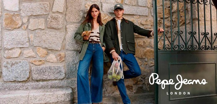 Una donna e un uomo in giacche casual e jeans si appoggiano a un muro di pietra, tenendo del pane e una borsa della spesa all'aperto vicino a un cancello nero.