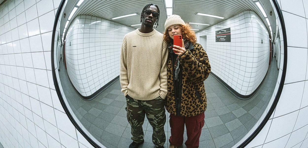Pyöreä peili heijastaa kahta henkilöä laatoitetussa käytävässä. Toinen pitää päällään beigeä ribbineulepuseroa ja maastokuvioisia housuja; toinen ruskeaa leopardi-kuvioista takkia.