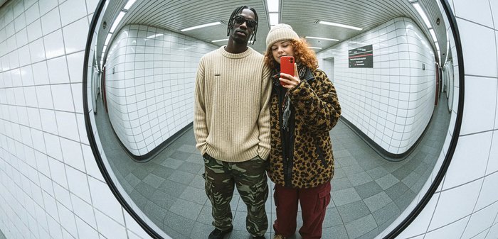 Ένας στρογγυλός καθρέφτης αντανακλά δύο άτομα σε έναν διάδρομο με πλακάκια. Ο ένας φοράει ένα μπεζ ριγωτό πουλόβερ και παντελόνι παραλλαγής· ο άλλος ένα καφέ παλτό με λεοπάρ εκτύπωση.