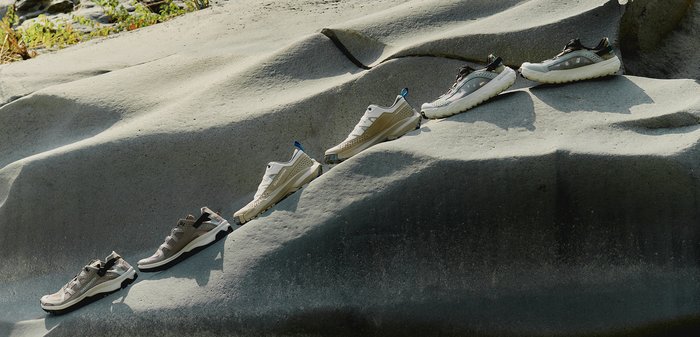 Three pairs of beige and brown hiking shoes placed on uneven gray rock surfaces outdoors under sunlight.