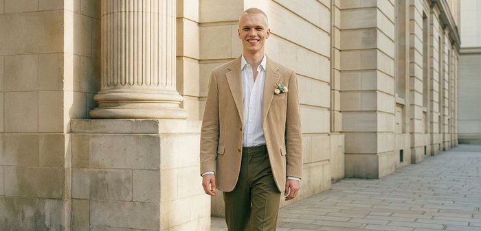 Άνδρας με μπεζ σακάκι και άσπρο πουκάμισο περπατά με αυτοπεποίθηση κατά μήκος πέτρινων κολόνων σε φωτεινό υπαίθριο περιβάλλον.