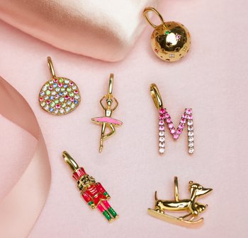 Le charm in oro includono un disco colorato di strass, una ballerina rosa, una "M" rosa, uno schiaccianoci e un cane d'oro su una slitta, tutti su tessuto rosa chiaro.