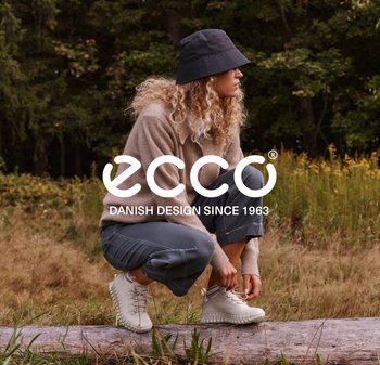 Woman with curly hair in casual clothes and bucket hat crouching outdoors tying white boots on a log in natural setting.