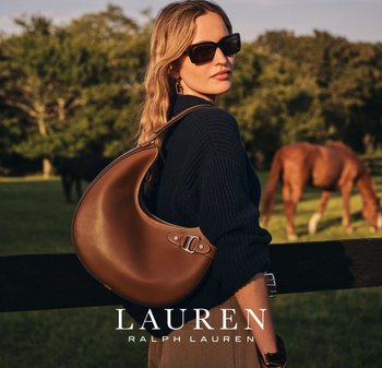Una donna con un maglione scuro e occhiali da sole, che porta una borsa a tracolla in pelle marrone, in piedi vicino a una recinzione in un campo con cavalli al pascolo.