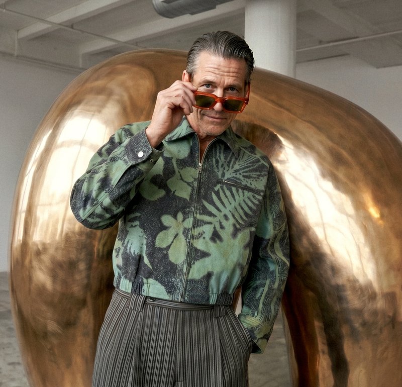 Uomo con giacca verde a fantasia e pantaloni a righe che si aggiusta occhiali da sole arancioni, davanti a una grande scultura astratta in bronzo lucido in uno spazio espositivo.