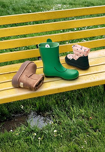 Três pares de calçado numa banca amarela: bota de borracha verde para a chuva, bota castanha peluda e bota preta de tornozelo com punho dobrado em padrão rosa.