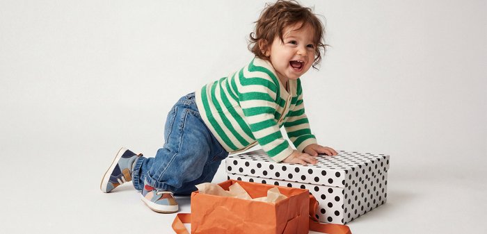 Νήπιο που φοράει πουλόβερ με πράσινες ρίγες και τζιν, γονατισμένο με τα χέρια πάνω σε ένα μεγάλο κουτί δώρου με πουά δίπλα σε μια πορτοκαλί τσάντα με χαρτί περιτυλίγματος.