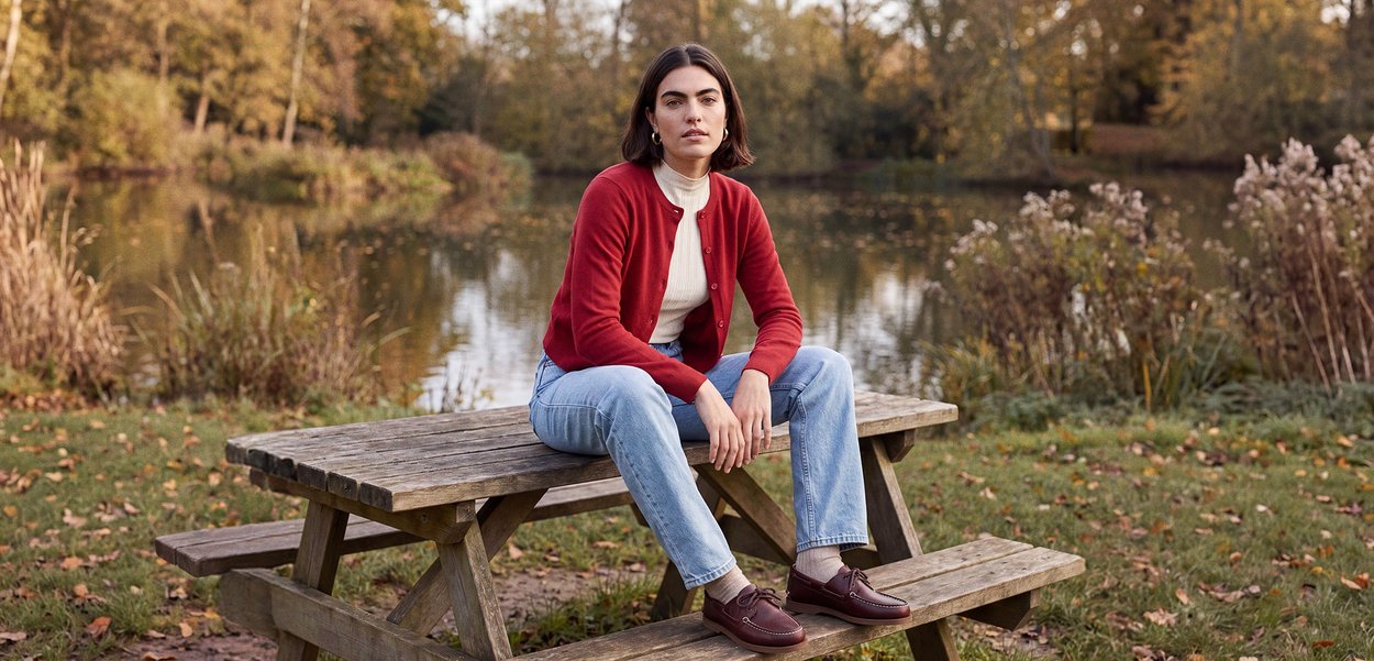 Una mujer con una chaqueta de punto roja y vaqueros está sentada en una mesa de pícnic de madera junto a un estanque en un parque otoñal.