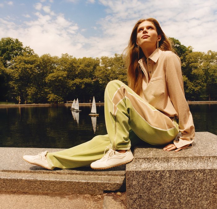 Camicia beige con bottoni e tasca, pantaloni della tuta verde e beige, e sneakers bianche. Modello seduto vicino all'acqua con barche a vela sullo sfondo.