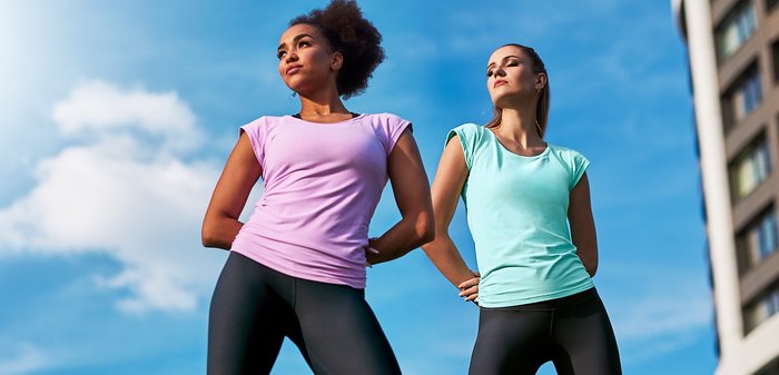 Twee vrouwen dragen nauwsluitende sporttops: één in lichtpaars, de ander in muntgroen, beide gecombineerd met zwarte leggings. Gladde, rekbare stof.