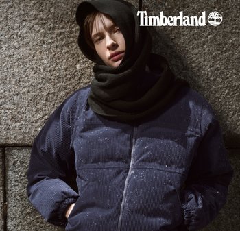 Marineblaue Cordjacke mit Steppung in Rippstruktur und elastischen Bündchen, kombiniert mit einem schwarzen Strickschal, vor einer Steinmauer. Timberland-Logo darüber.