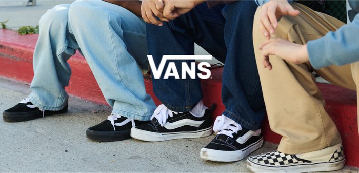 Tre adolescenti di diversa provenienza indossano scarpe Vans e sono seduti su un marciapiede, sorridendo e chiacchierando all'aperto vicino a un muro grigio e una scala.