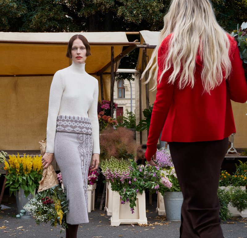 White knit turtleneck sweater paired with a patterned grey skirt. Flowers and plants in pots are arranged in the background.