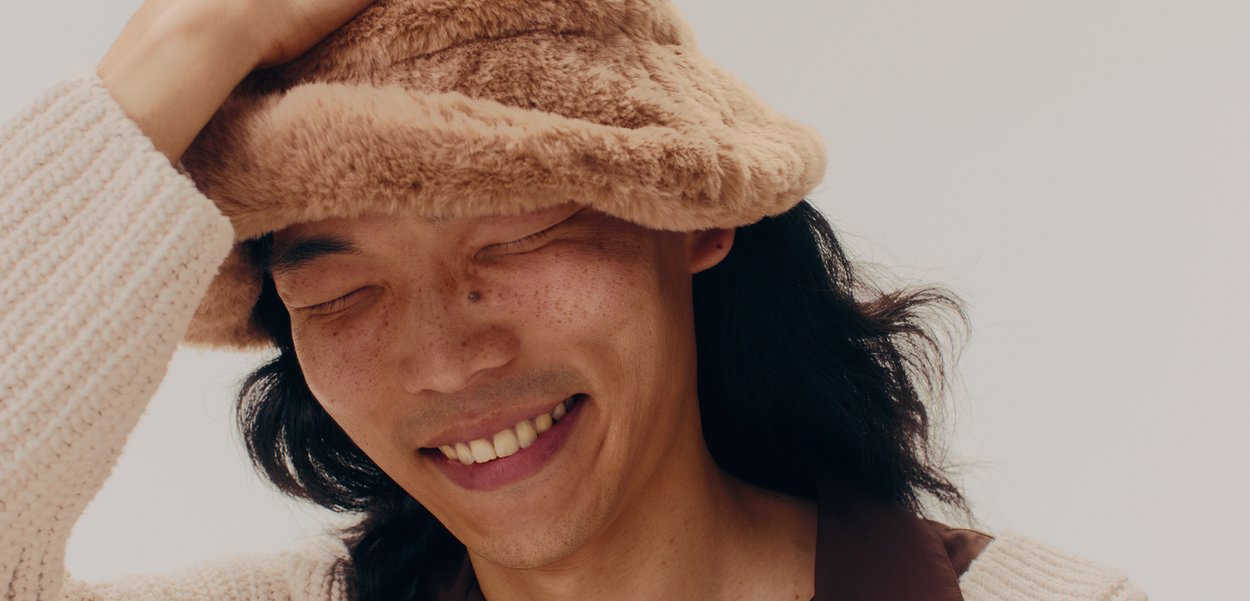 Pălărie tip bucket din blană artificială de culoare bej deschis, cu o textură moale și o margine lată și flască. Persoana are părul închis la culoare și poartă un pulover texturat de culoare crem.