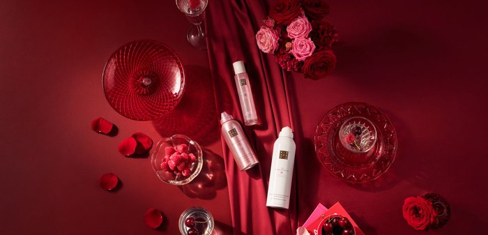Två sprayflaskor i rosa och vitt, en rund röd glasfat, en klar skål med hallon, rosenblad och blommor på en röd bakgrund.