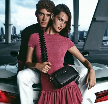 Jeune homme en pull noir assis sur le coffre d’une voiture blanche derrière une femme en robe rose tenant un sac bandoulière noir dans un cadre urbain moderne.