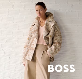 Vrouw draagt beige wijde broek, lichtroze trui en beige jas met patroon, staand tegen een witte bakstenen muur met de tekst "BOSS".