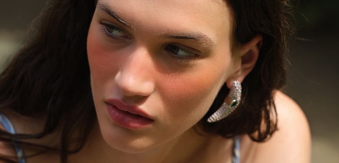 Jeune femme aux longs cheveux foncés regarde sur le côté, portant de grandes boucles d'oreilles en argent et un maquillage naturel avec un blush pêche.