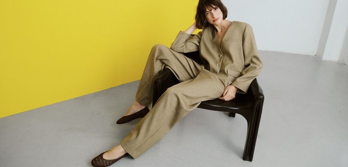 Persoon gekleed in een beige maatpak met broek met wijde pijpen, die een donkerbruine leren draagtas vasthoudt, staand naast een moderne metalen stoel.