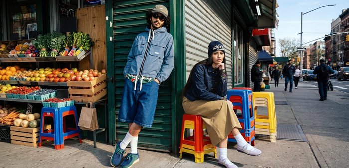 Deux jeunes adultes en tenue décontractée urbaine s'appuient et s'assoient sur des tabourets colorés devant un marché de produits frais sur un trottoir en ville.