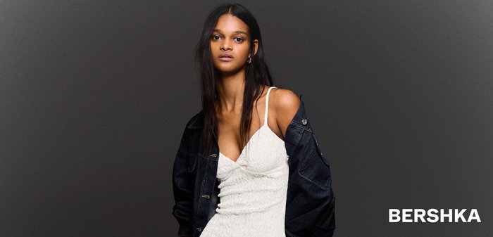 Femeie îmbrăcată într-o rochie slip texturată albă cu bretele subțiri și o jachetă din denim închis drapată peste umeri, pe un fundal gri.