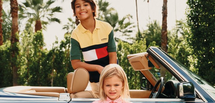 Junge in einem mehrfarbigen Poloshirt lehnt aus einem blauen Cabriolet, Mädchen in einem rosa gestreiften Kleid steht davor, mit Palmen im Hintergrund.