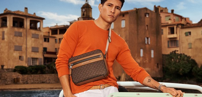 Mann in orangefarbenem Pullover mit einer Umhängetasche steht am Wasser, im Hintergrund sind alte europäische Gebäude bei teilweise bewölktem Himmel zu sehen.