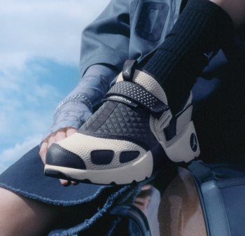 Personne portant des baskets Jordan noires et beiges avec des chaussettes noires, assise sur une surface métallique bleue sous un ciel partiellement nuageux.