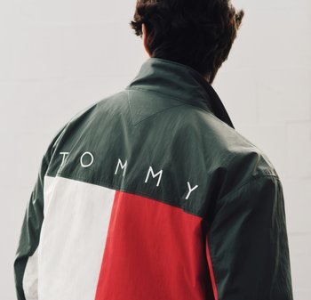 Persoon draagt een groene, witte en rode jas met de tekst "TOMMY" op de achterkant, met de rug naar de kijker, tegen een neutrale, lichte achtergrond.