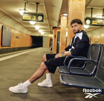 Hombre vestido con ropa deportiva en blanco y negro y zapatillas blancas Reebok sentado en un banco de metal en una estación de metro vacía.